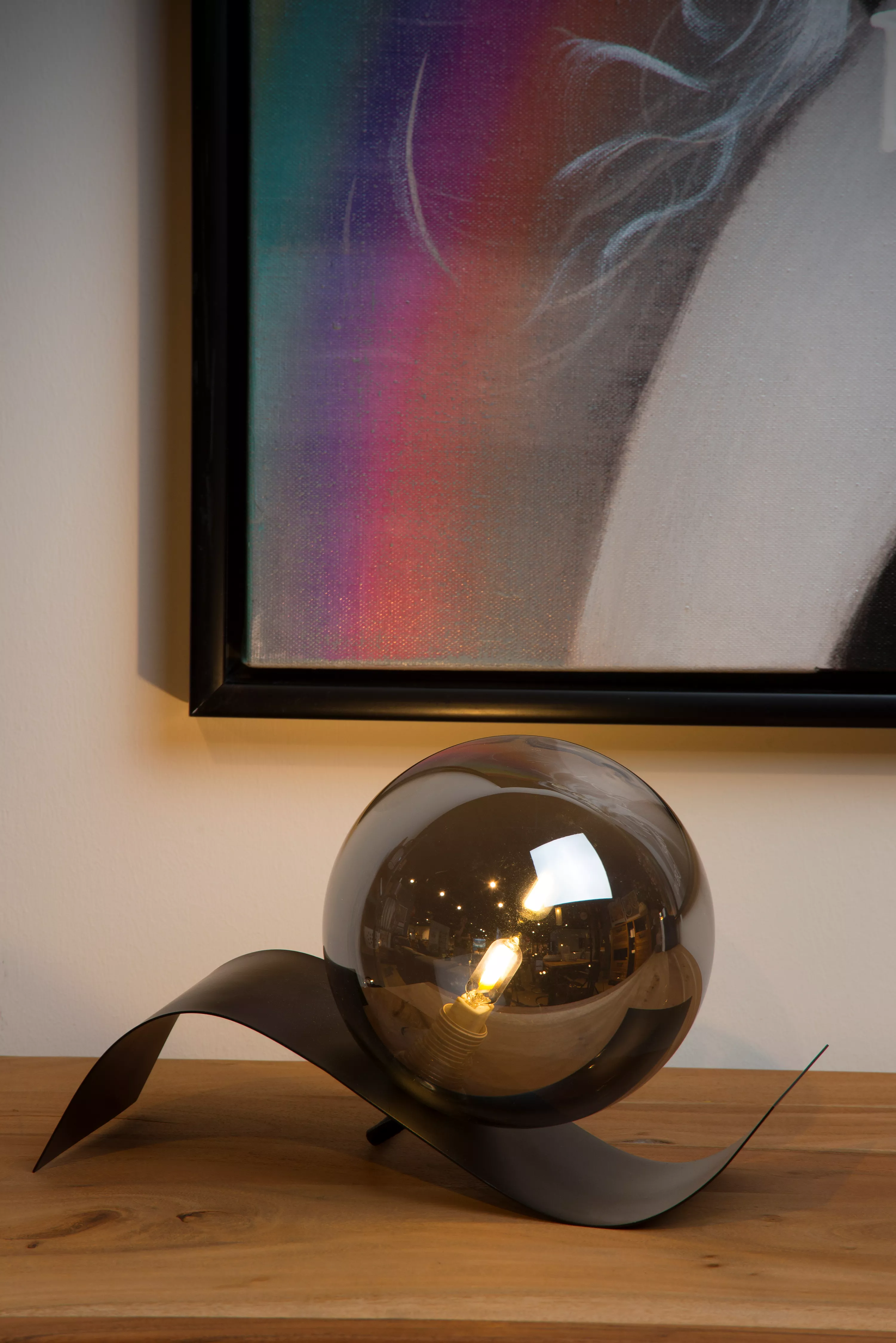 Stolová lampička Yoni so zvlnenou kovovou základňou a guľou z dymového skla vydáva v obývačke alebo spálni príjemné rozptýlené svetlo.