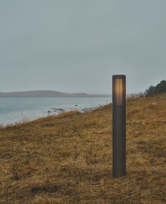 Vonkajší záhradný svetelný stĺpik Aludra 95 Seaside od Nordluxu v modernom minimalistickom dizajne. Vďaka svojmu špecifickému tvaru vytvára na okolí hru svetla a tieňa. Vyrobený z odolného materiálu, dostupný v troch farbách – čierna, antracitová, metalická hnedá.